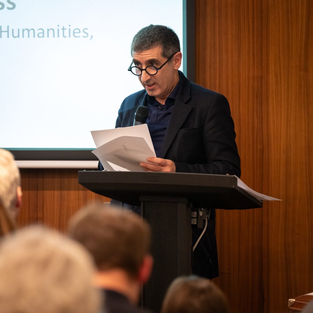 Speaker in a dark blazer reading from papers at a podium, with an audience visible in the foreground.