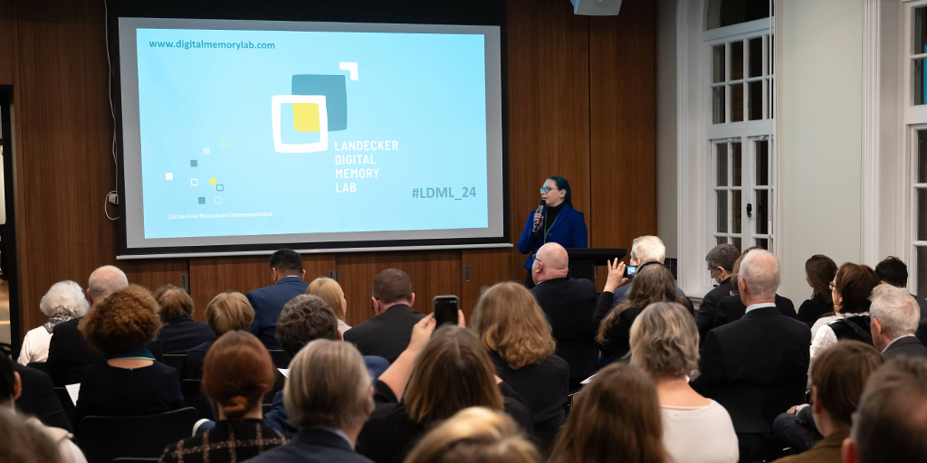 A woman in blue jacket standing in front of a bunch of sitting people, giving a speech.