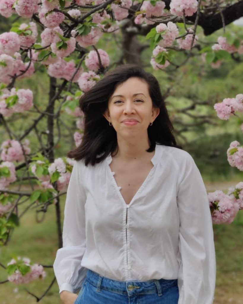 A person stands outdoors, smiling softly amid blooming pink cherry blossoms. They are wearing a white blouse and blue jeans, with one hand in their pocket, creating a relaxed and natural vibe. The lush, floral background adds a sense of tranquillity and beauty to the scene, enhancing the warmth of their expression.