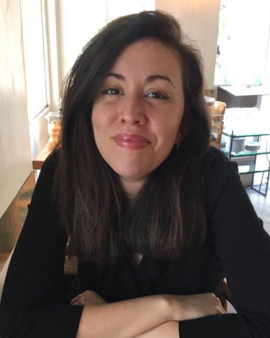 A person with long dark hair smiles warmly while seated at a table in a cozy, well-lit café. They are leaning forward with their arms crossed on the table, creating a relaxed and approachable vibe. The background shows wooden chairs and a softly lit interior, adding to the inviting atmosphere of the scene.