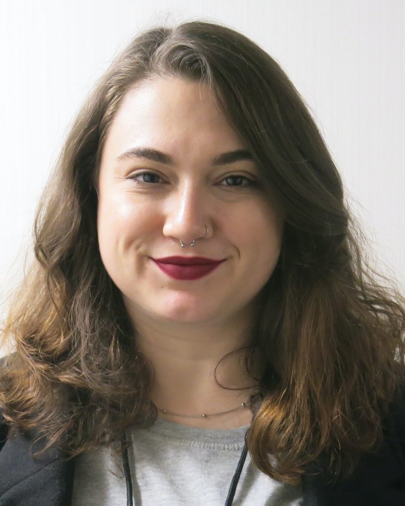 A person with wavy dark hair smiles gently in this close-up portrait. They have a septum piercing and wear bold red lipstick, adding character to their look. Dressed in a casual top and blazer, they exude a friendly and approachable vibe, with a simple background keeping the focus on their expressive features.