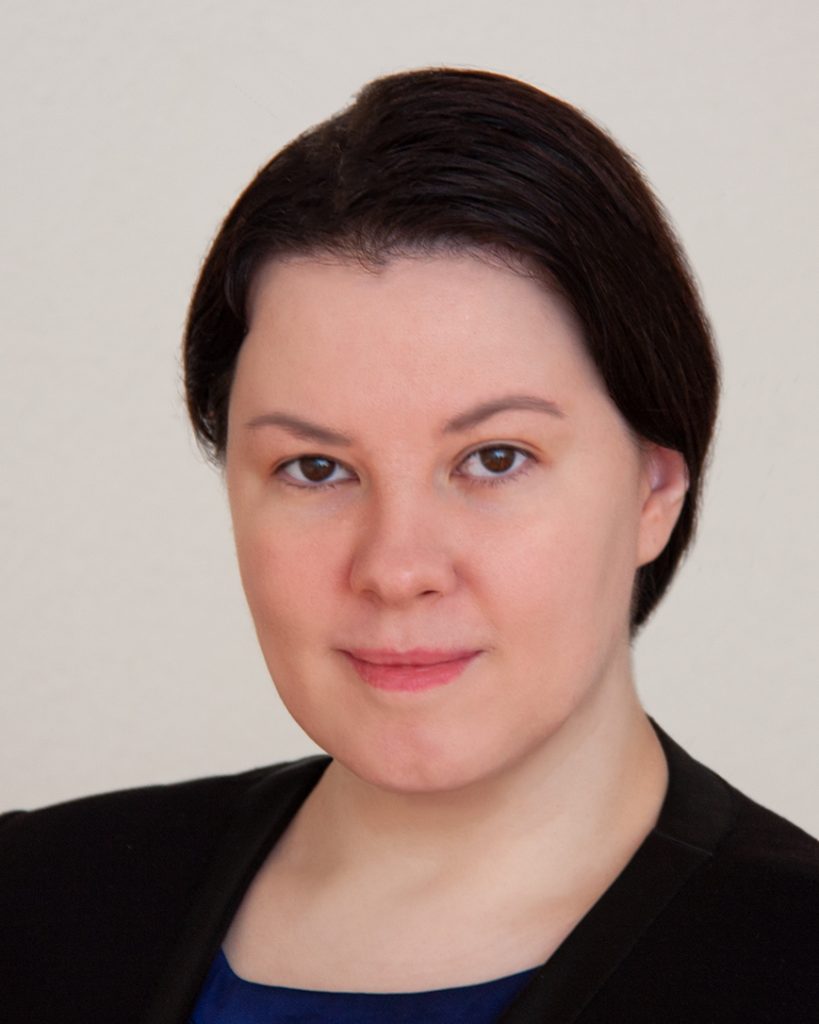 A person with neatly styled dark hair is shown in a close-up portrait. They have a calm, confident expression, wearing a dark blazer over a blue top. The plain, light background keeps the focus on their face, emphasizing their poised and professional appearance.