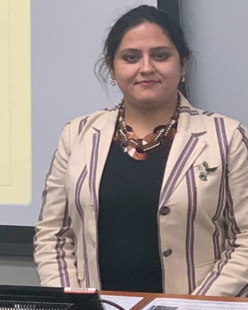 A person stands confidently at a lectern, wearing a striped blazer over a dark top, accessorized with a statement necklace and a brooch. They have a composed expression, with their hair pulled back, presenting a professional and poised demeanor against the background of a presentation screen.