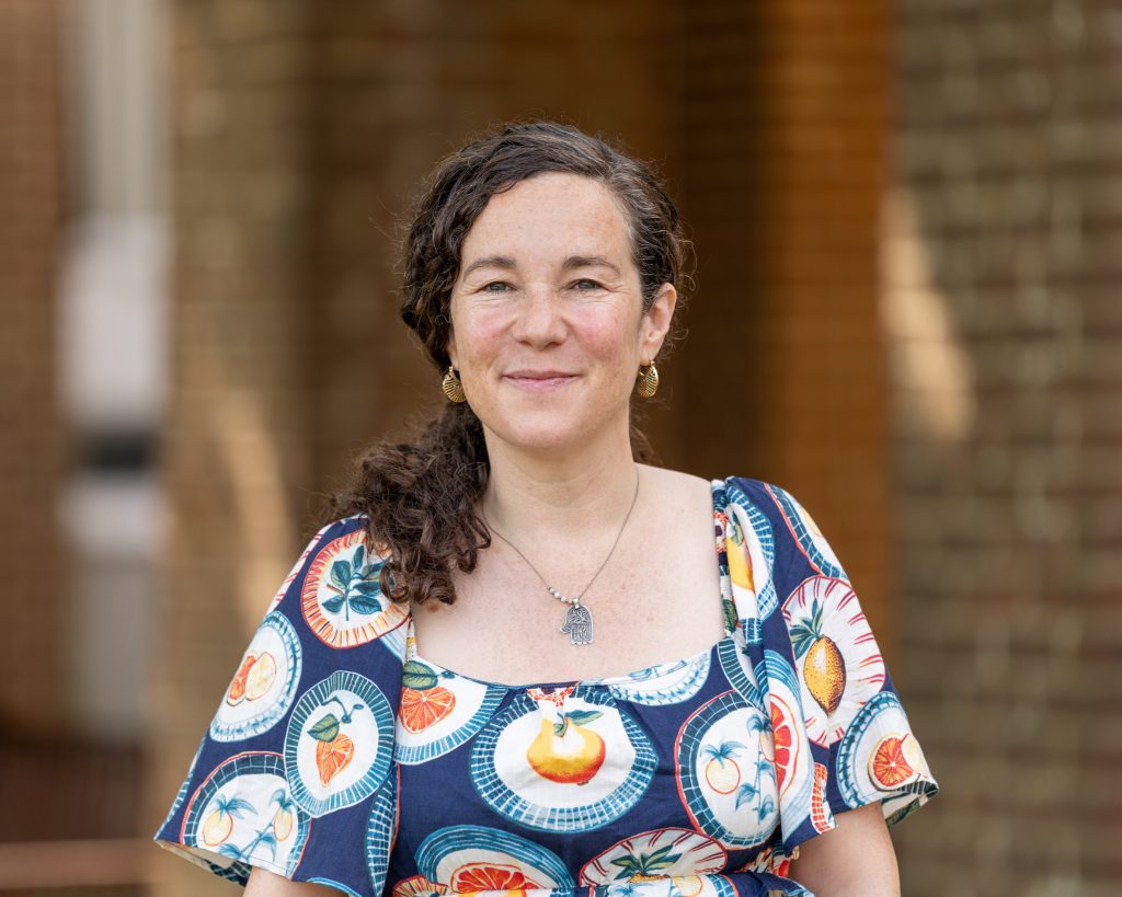 A person with curly dark hair stands outdoors in front of a brick building, wearing a colorful patterned dress featuring fruit designs. She has a warm expression, with soft, natural light enhancing the relaxed atmosphere. Her hair is pulled to one side, and she wears subtle jewelry, including a necklace and earrings.