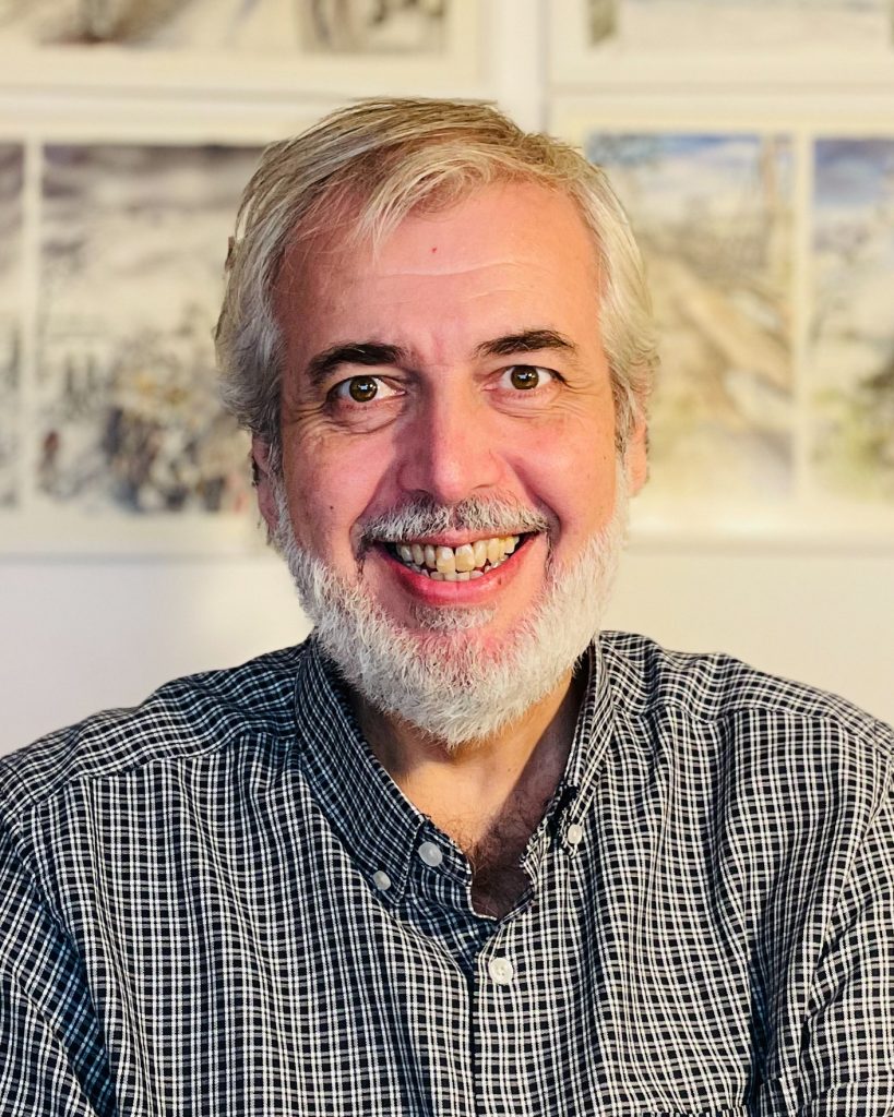 A person with a warm smile and a neatly trimmed white beard is shown in a close-up portrait. They are wearing a checkered shirt and sitting against a backdrop of framed artwork, adding a creative and inviting touch to the scene. Their cheerful expression and relaxed posture convey friendliness and approachability.