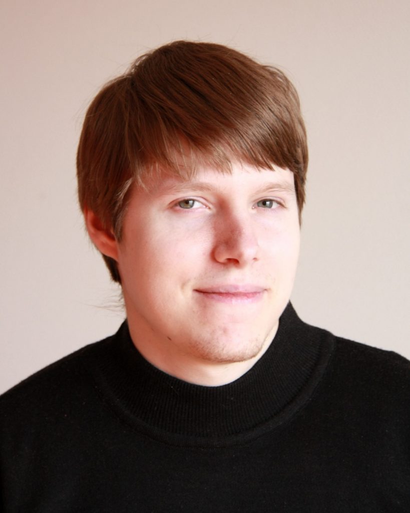 A person with short, light brown hair is shown in a close-up portrait against a plain background. They are wearing a dark turtleneck sweater and have a calm, gentle expression. The minimalist setting keeps the focus on their face, conveying a composed and approachable demeanour.