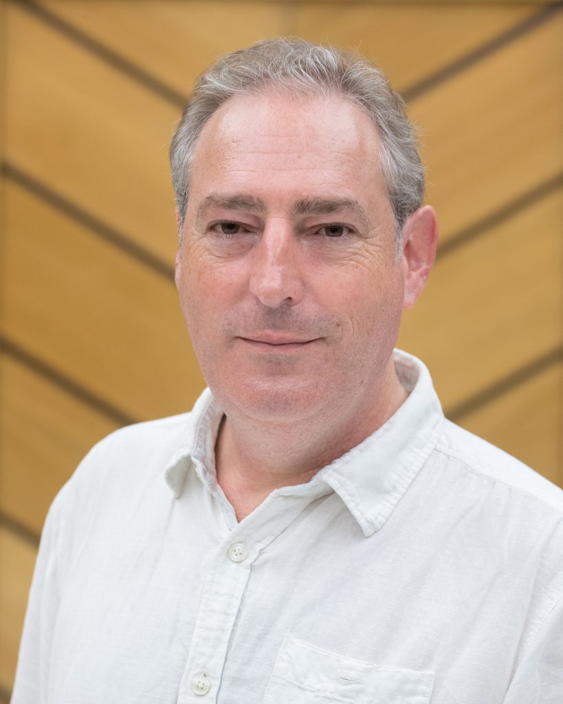 This image shows a man with short, light gray hair, wearing a light-colored button-up shirt. He has a calm expression, and the background consists of a warm-toned, geometrically patterned wall, providing a professional and composed setting.