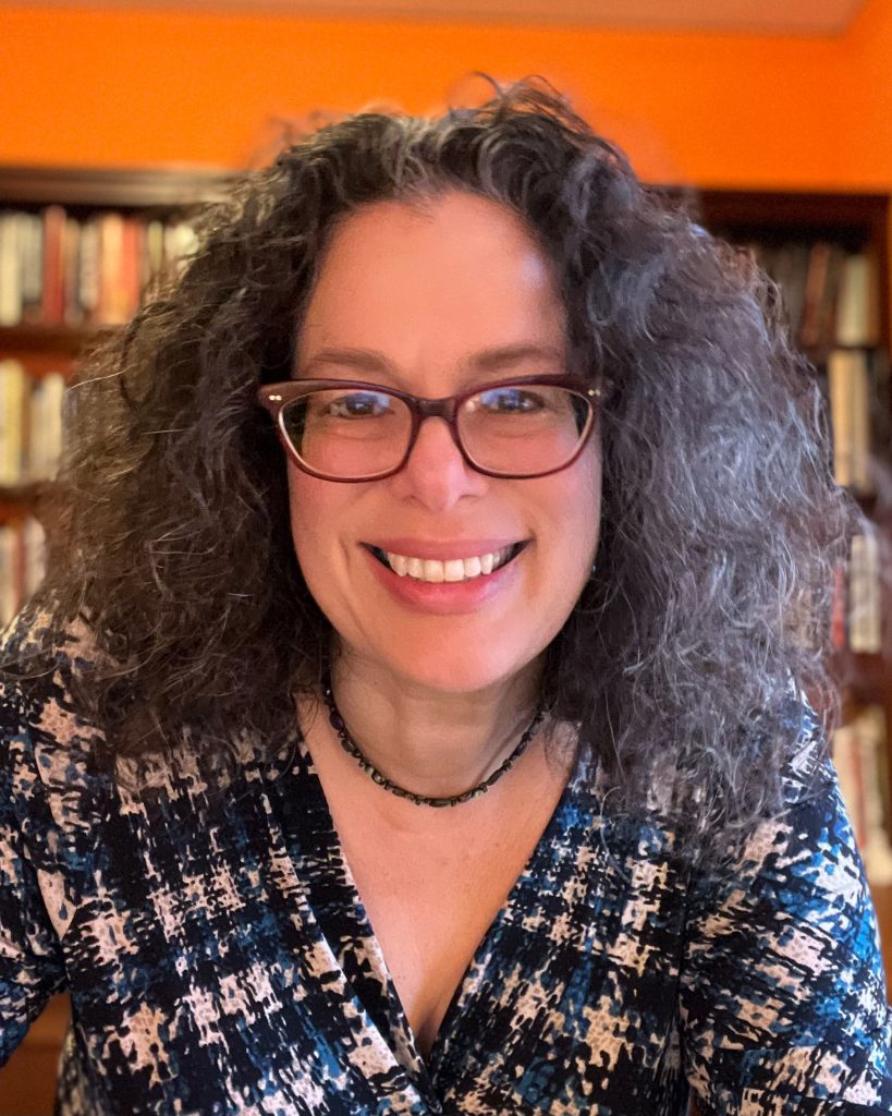 A person with curly grey hair and glasses smiles warmly in a cozy room with bookshelves in the background. Dressed in a patterned blouse and wearing a simple necklace, they exude a friendly and approachable vibe. The orange walls and books add a touch of warmth and personality to the setting.