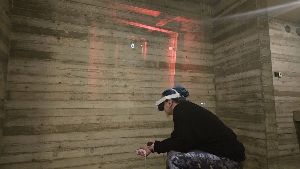 A woman with blue hair uses a VR headset and hand controllers in a wooden room.