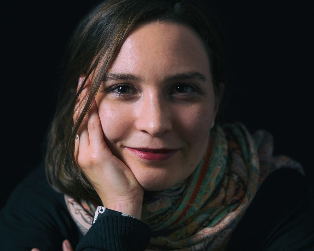 A woman with a thoughtful expression, resting her head on her hand. She has a warm smile, dark hair, and is wearing a colorful scarf. The background is dark, which highlights her face and gives the portrait an intimate feel.