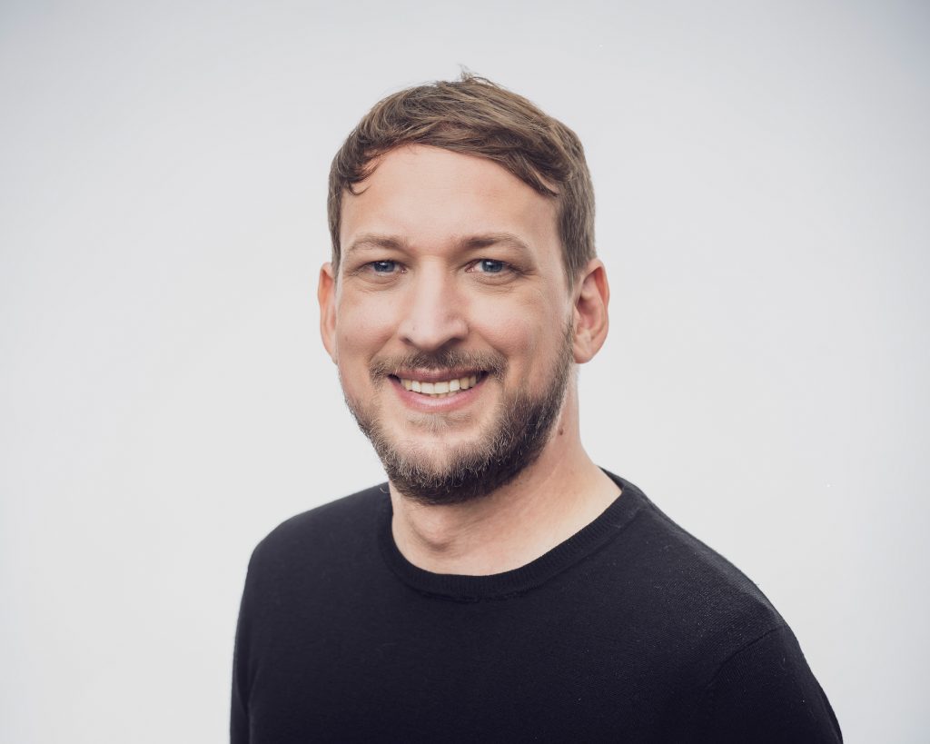 The photo shows an individual with short hair and a trimmed beard, wearing a black sweater. They have a friendly expression and a slight smile, creating a warm and approachable look. The background is a neutral, light grey, which brings focus to their face and adds a professional tone to the portrait.