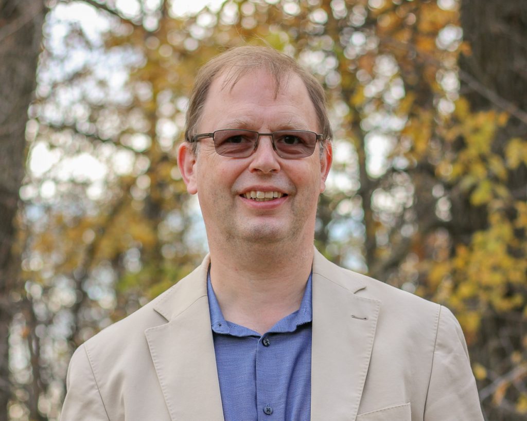 A person stands outdoors in a natural setting with autumn foliage in the background. They are wearing glasses, a beige blazer, and a blue collared shirt, smiling warmly at the camera. The fall colours and soft lighting create a friendly and approachable atmosphere.