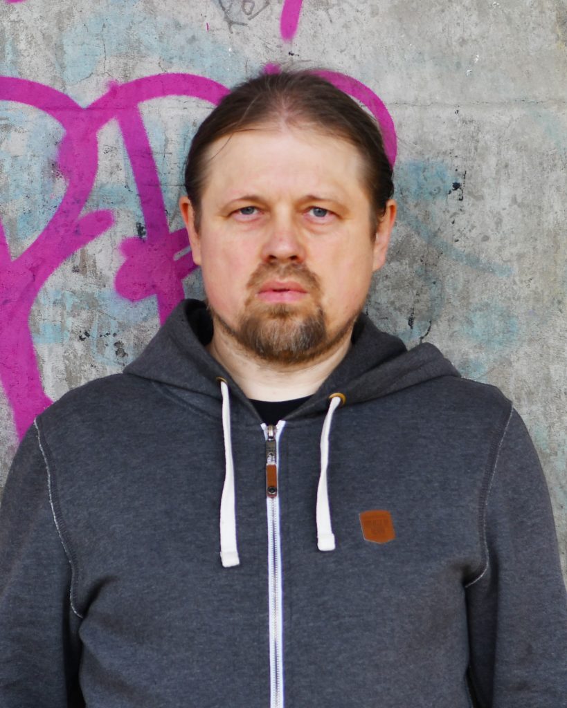 A person with a serious expression stands in front of a concrete wall with pink graffiti. They are wearing a dark hoodie with white drawstrings, giving a casual and urban feel to the portrait. The background and their focused look add an edgy, introspective vibe to the image.