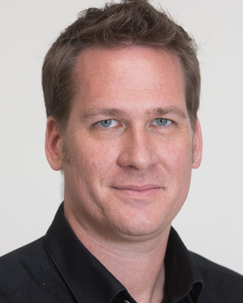 A person with short hair and a calm expression is shown in a close-up portrait against a plain background. They are wearing a black collared shirt, giving them a professional appearance. Their direct gaze and slight smile convey a confident and approachable presence.