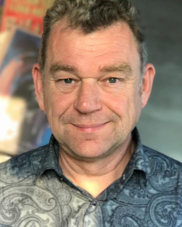 This image shows a man with short, curly hair, wearing a patterned shirt. He has a slight smile and appears to be in an indoor setting with a blurred background. The overall tone is casual and warm.