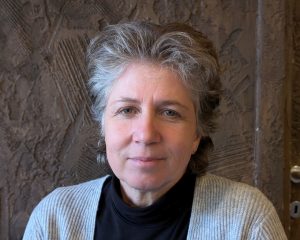 This image shows a woman with short, wavy gray hair, wearing a black top with a light gray cardigan. She has a calm expression and is seated in front of a textured, brown wall. The setting has a warm, natural ambiance.