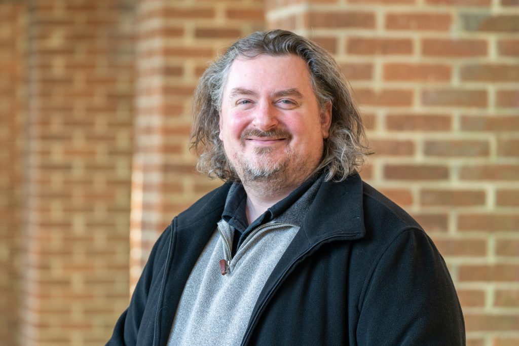 A man with shoulder-length hair smiles at the camera against a brick backdrop.