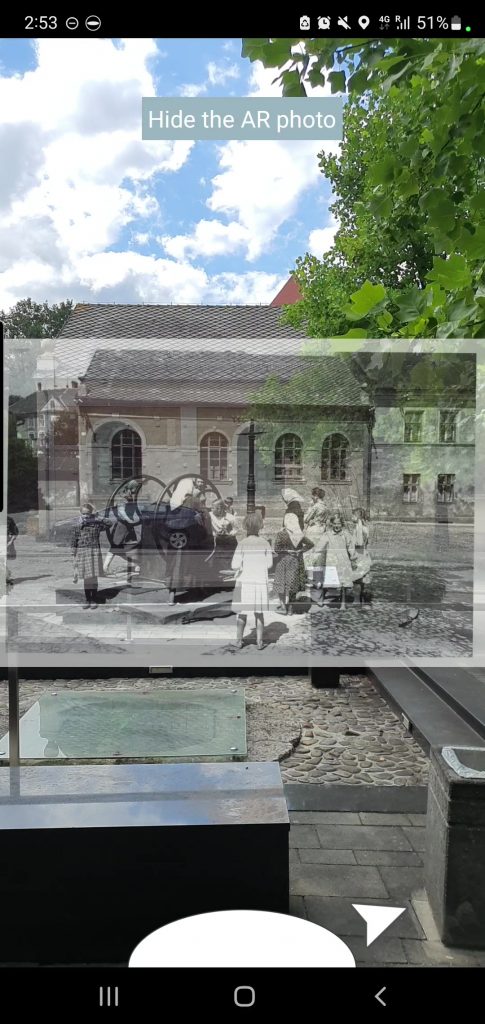 Augmented reality view of a historical photograph overlaid on a modern setting. The AR image shows a group of people gathered around a fountain, blending with a contemporary outdoor scene of trees, a building, and cobblestone paving.