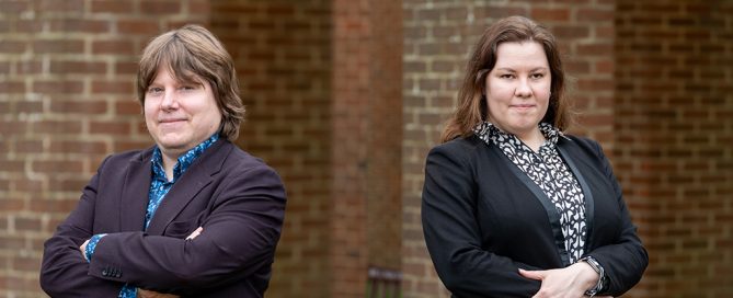 A professional portrait of two individuals standing against a brick background, both dressed in business attire with arms crossed, looking confidently at the camera.