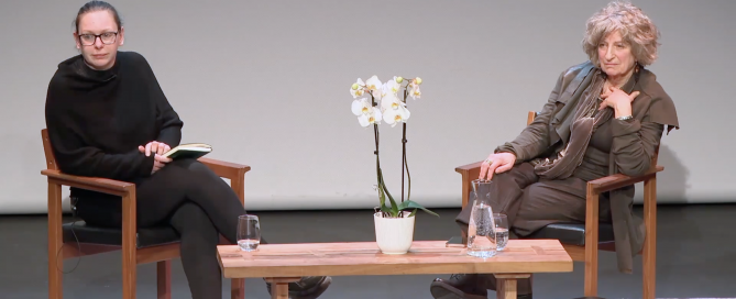 A staged conversation between two women seated on wooden chairs with a small table between them, featuring a potted white orchid, water glasses, and a jug. The setting appears to be a formal discussion or interview.