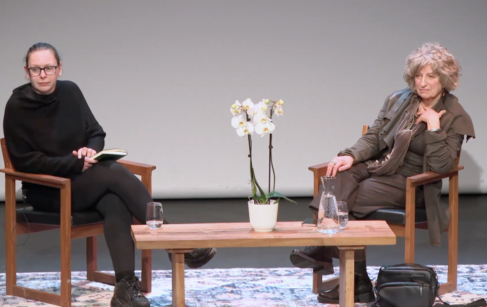 A staged conversation between two women seated on wooden chairs with a small table between them, featuring a potted white orchid, water glasses, and a jug. The setting appears to be a formal discussion or interview.