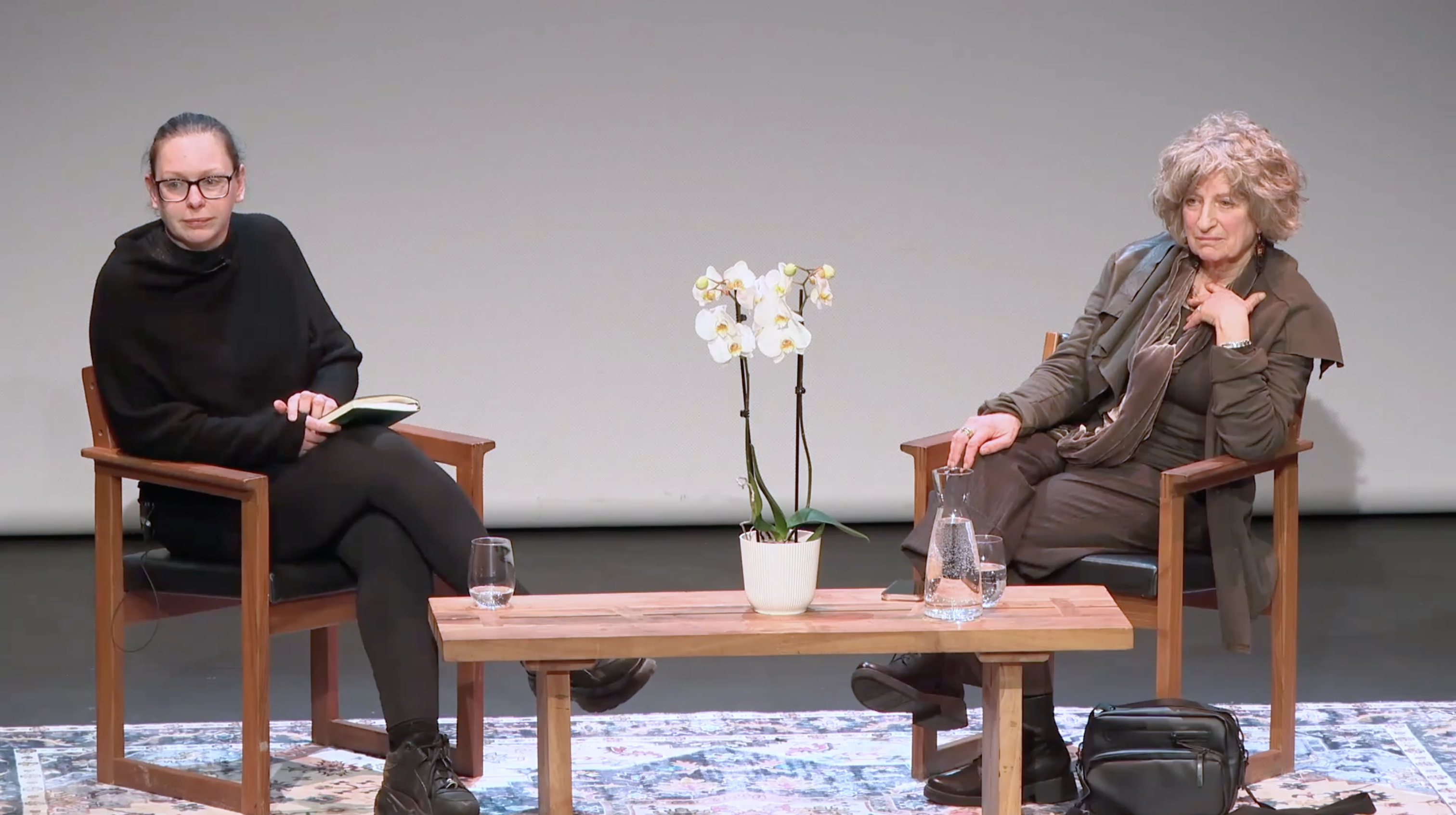 A staged conversation between two women seated on wooden chairs with a small table between them, featuring a potted white orchid, water glasses, and a jug. The setting appears to be a formal discussion or interview.